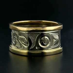 May include: A wide gold band ring with a detailed carved design in silver. The design features a stylized animal or creature with a circular eye.