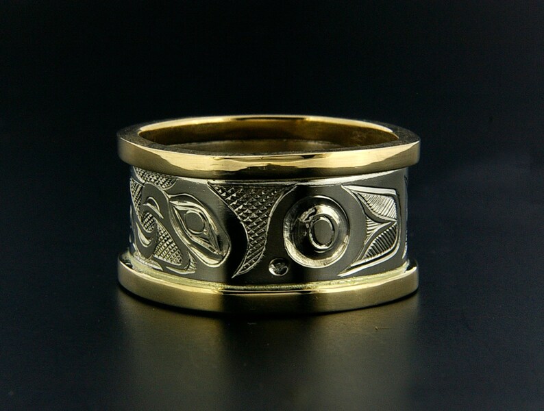 May include: A wide gold band ring with a detailed carved design in silver. The design features a stylized animal or creature with a circular eye.