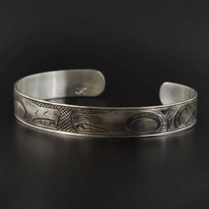 May include: A silver bracelet featuring an intricate design of two stylised animal faces. The bracelet is crafted from polished metal and has a smooth, rounded surface.