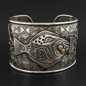 Può includere: Un braccialetto in argento con un motivo geometrico e un design a rilievo di un pesce con accenti dorati.