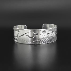 Op de afbeelding: Een zilveren manchetarmband met een gegraveerd ontwerp. De armband heeft een gestileerde afbeelding van een walvis of een ander zeedier. Het ontwerp omvat een oog, vinnen en andere details. De armband wordt op een donkere ondergrond getoond.
