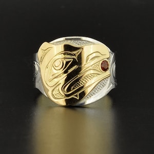 Puede incluir: Un anillo de plata y oro con un diseño tallado de la cara de un oso. El ojo del oso es una piedra preciosa roja.