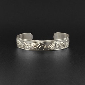 May include: A silver bracelet with an intricate design of a whale and waves. The bracelet is made of polished metal and has a smooth finish.