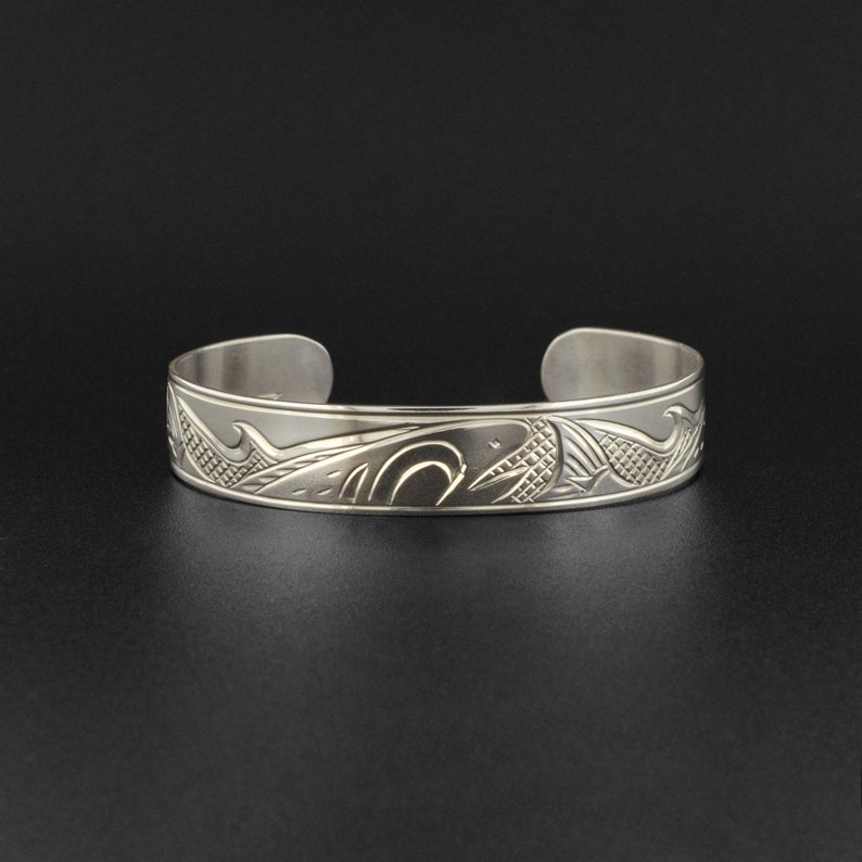 May include: A silver bracelet with an intricate design of a whale and waves. The bracelet is made of polished metal and has a smooth finish.