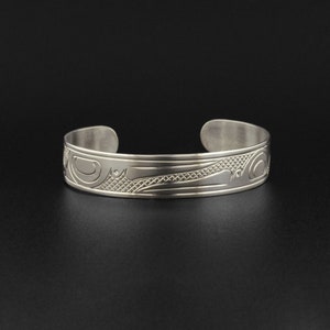 May include: A silver bracelet with a detailed design of a stylized bird or animal. The design is engraved into the metal and has a textured finish.