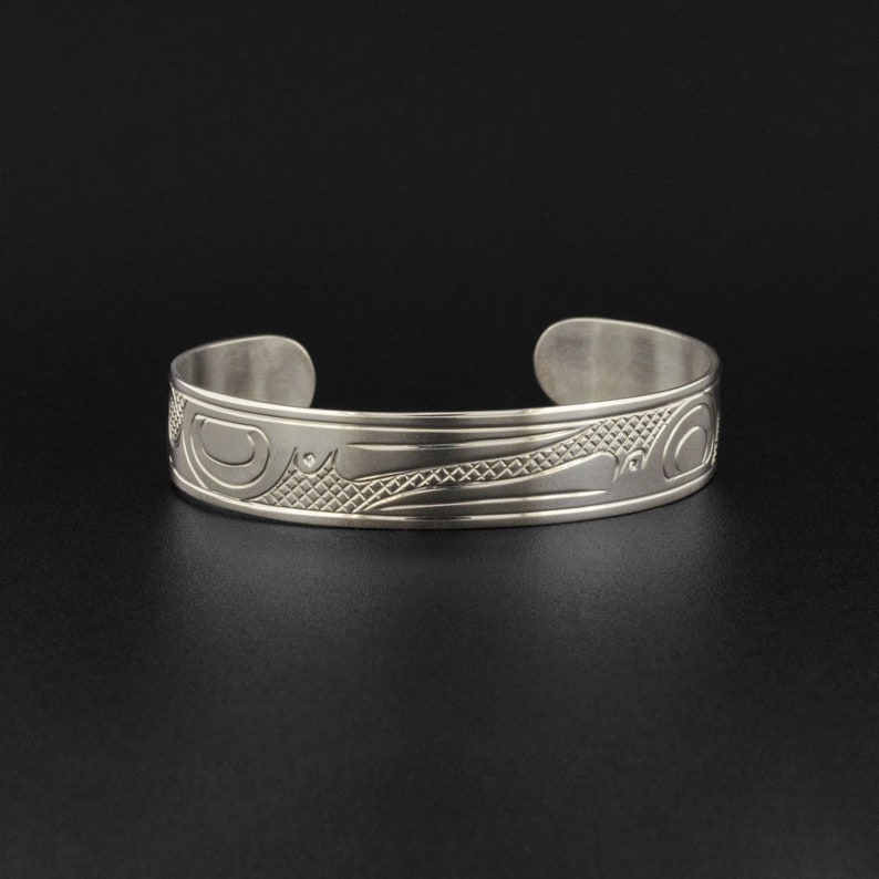 May include: A silver bracelet with a detailed design of a stylized bird or animal. The design is engraved into the metal and has a textured finish.
