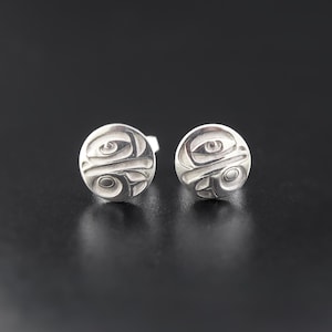 May include: A pair of silver stud earrings featuring a circular design with a stylized face, possibly a bird or animal, in a Northwest Coast Indigenous art style.