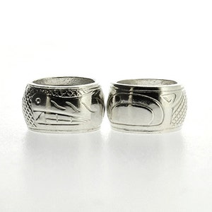 Könnte beinhalten: Zwei silberne Ringe mit aufwendigen Schnitzereien von indianischen Designs. Die Ringe liegen nebeneinander und sind beide zur Kamera gerichtet.