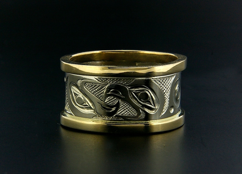 May include: A wide gold band ring with a detailed design of stylized animal eyes and feathers. The ring is made of two different metals, with a gold band around the edge.