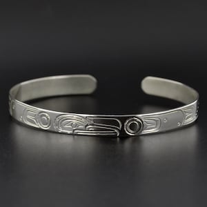 Puede incluir: Una pulsera de plata con un diseño tallado de un cuervo, una forma de arte tradicional de las Primeras Naciones de la costa noroeste.
