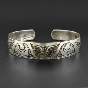 Könnte beinhalten: Ein silbernes Armband mit einem detaillierten Design eines stilisierten Vogels, möglicherweise ein Rabe oder Adler, mit komplizierten Linien und Mustern. Das Armband ist aus poliertem Metall gefertigt und hat eine glatte, abgerundete Oberfläche.
