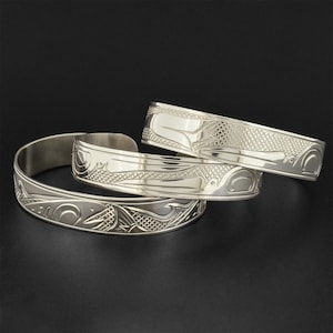 May include: Three silver bracelets with intricate carvings of stylized animals and geometric patterns. The bracelets are stacked on top of each other.