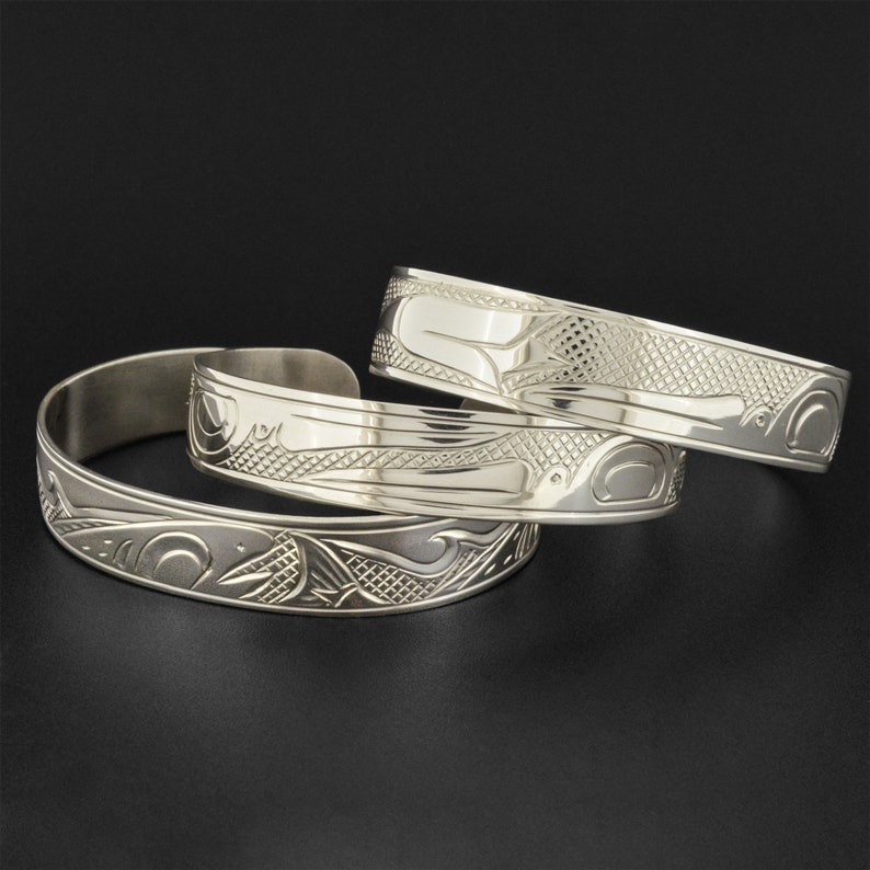 May include: Three silver bracelets with intricate carvings of stylized animals and geometric patterns. The bracelets are stacked on top of each other.