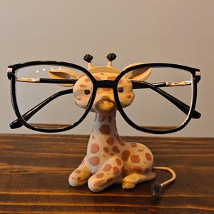 May include: A whimsical eyeglasses holder shaped like a giraffe, with a cream and brown spotted pattern. The giraffe holds a pair of black-framed glasses with gold accents. The holder sits on a dark wooden surface.