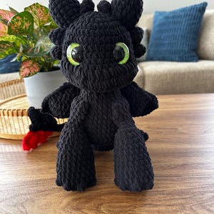 Può includere: Un peluche a forma di drago nero fatto a mano con grandi occhi verde brillante. Il drago ha piccole ali e una coda rossa e nera. È appoggiato su una superficie di legno.