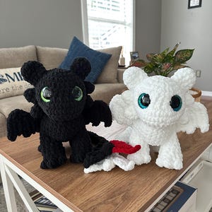 Può includere: Due peluche a forma di drago all'uncinetto, uno nero e uno bianco, con grandi occhi rotondi, verdi e blu. Il drago nero ha la punta della coda rossa. Sono esposti su un tavolo di legno.
