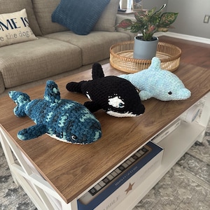 Puede incluir: Tres criaturas marinas de peluche de ganchillo: un tiburón azul y verde, una orca negra y blanca y un delfín azul claro. Se exhiben sobre una mesa de centro de madera con base blanca. La mesa también contiene un juego de Scrabble.