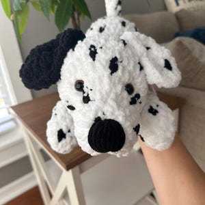 May include: A handmade white and black crochet Dalmatian dog. The plush toy has black spots, ears, and nose. The dog is lying down on a white table with a brown top.