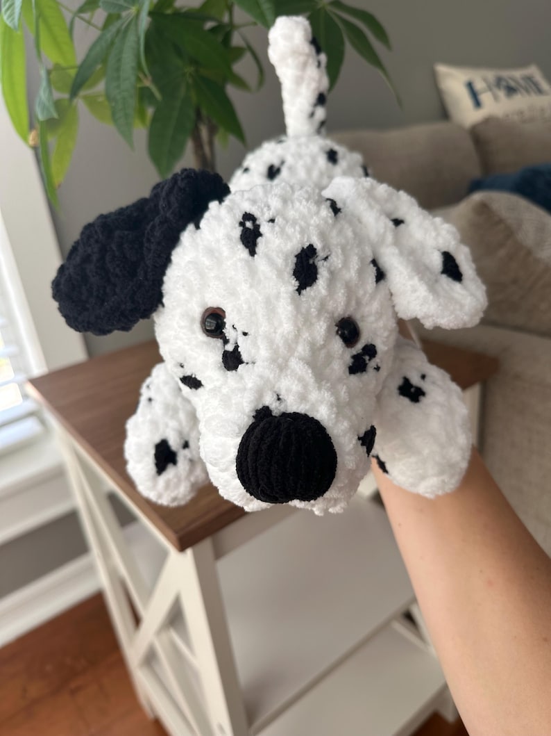 May include: A handmade white and black crochet Dalmatian dog. The plush toy has black spots, ears, and nose. The dog is lying down on a white table with a brown top.