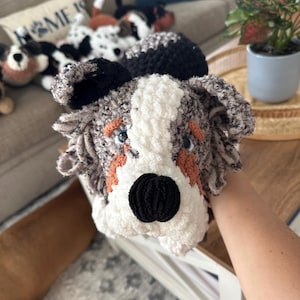 May include: A handmade crochet dog toy with a black nose, blue eyes, and orange accents. The dog's face is white and gray, with black ears. Other similar dog toys are visible in the background, suggesting a collection of handmade plush toys.