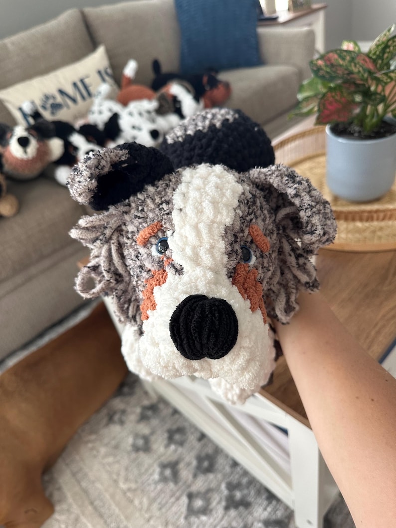 May include: A handmade crochet dog toy with a black nose, blue eyes, and orange accents. The dog's face is white and gray, with black ears. Other similar dog toys are visible in the background, suggesting a collection of handmade plush toys.