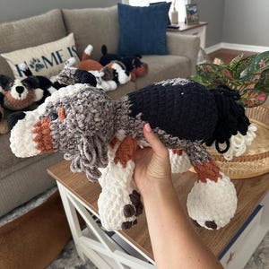 May include: A handmade crochet dog plushie in shades of gray, black, white, and brown. The dog has floppy ears, a fluffy tail, and brown spots on its paws. Other similar plushies are visible in the background, suggesting a collection of dog-themed items.