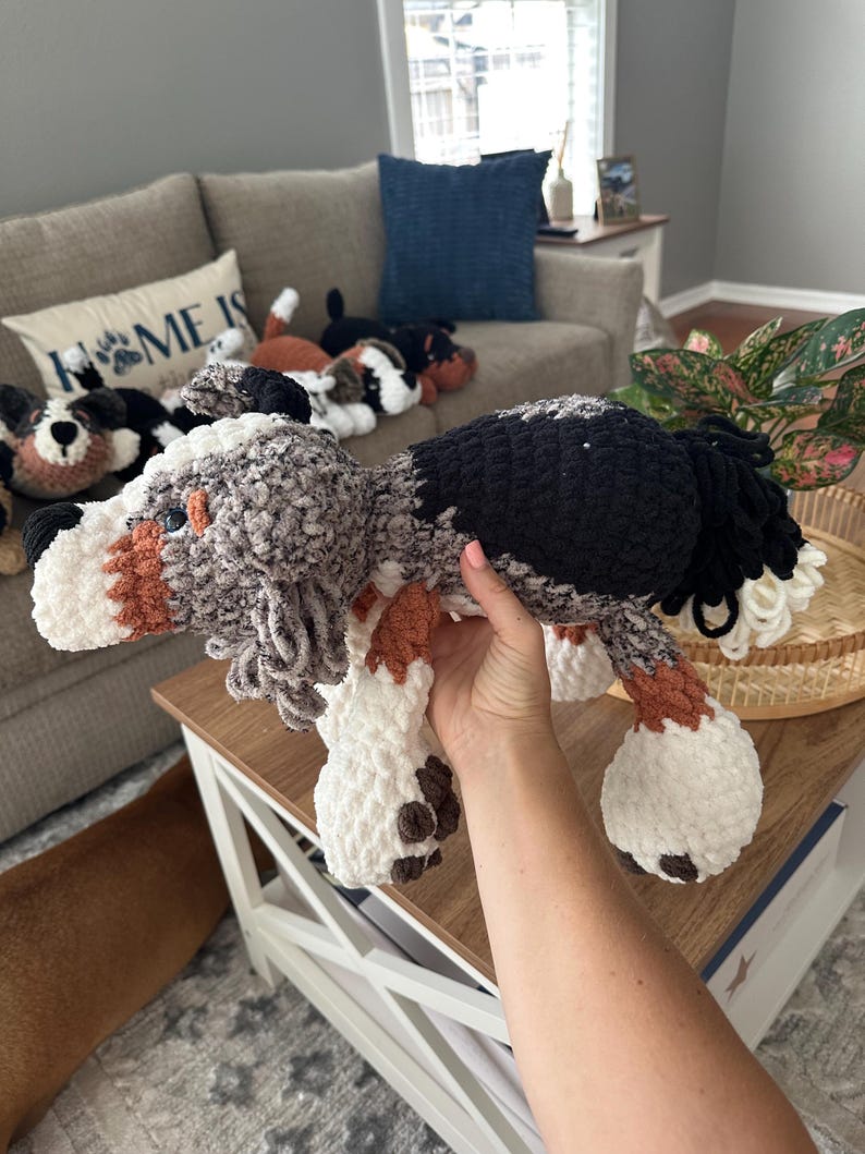 May include: A handmade crochet dog plushie in shades of gray, black, white, and brown. The dog has floppy ears, a fluffy tail, and brown spots on its paws. Other similar plushies are visible in the background, suggesting a collection of dog-themed items.