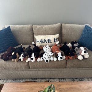 May include: A collection of handmade crochet dog plushies in various colors and patterns, including black, brown, white, and dalmatian-style spots. They are arranged on a beige couch with a decorative pillow that reads "HOME IS where the dog is."