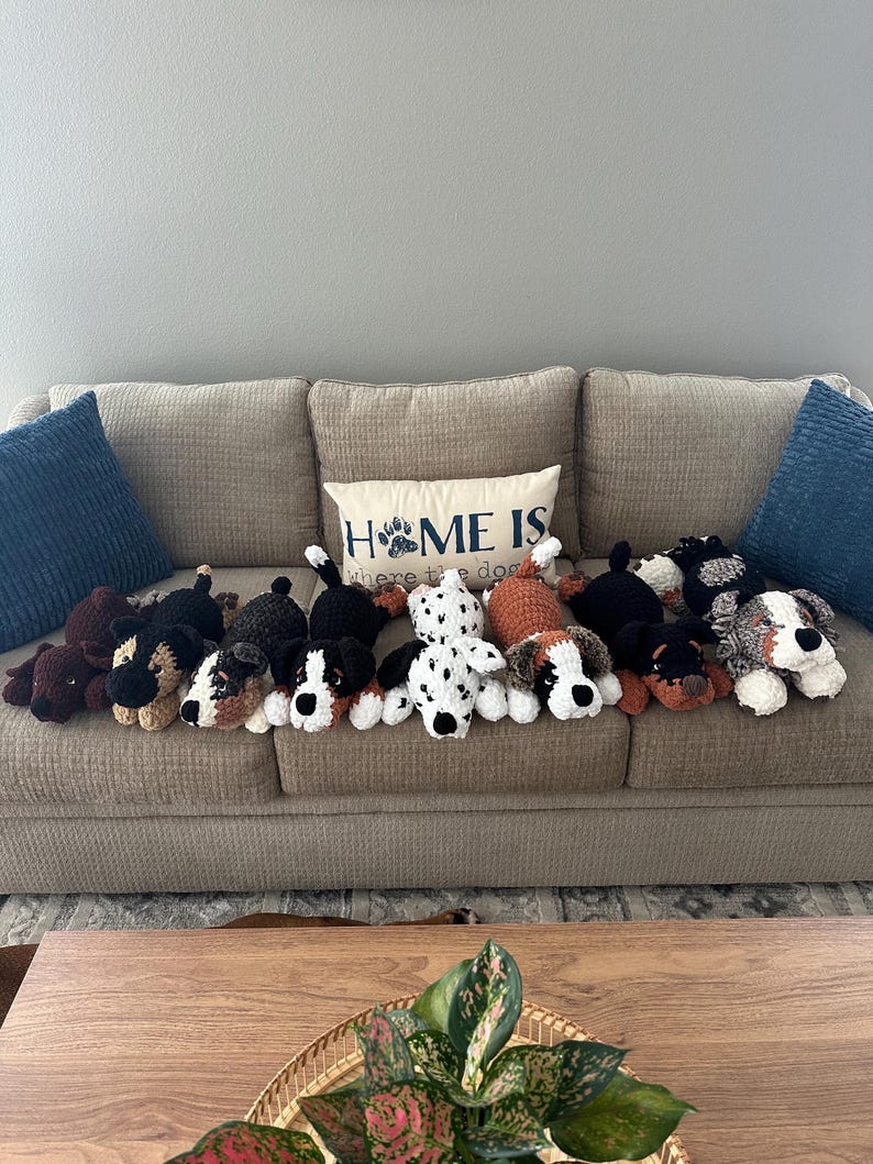May include: A collection of handmade crochet dog plushies in various colors and patterns, including black, brown, white, and dalmatian-style spots. They are arranged on a beige couch with a decorative pillow that reads "HOME IS where the dog is."