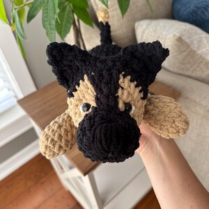 May include: A handmade crochet dog toy in the style of a German Shepherd. The dog has a black snout, ears, and back, with tan and cream-colored accents. The toy is lying down, with its paws extended.