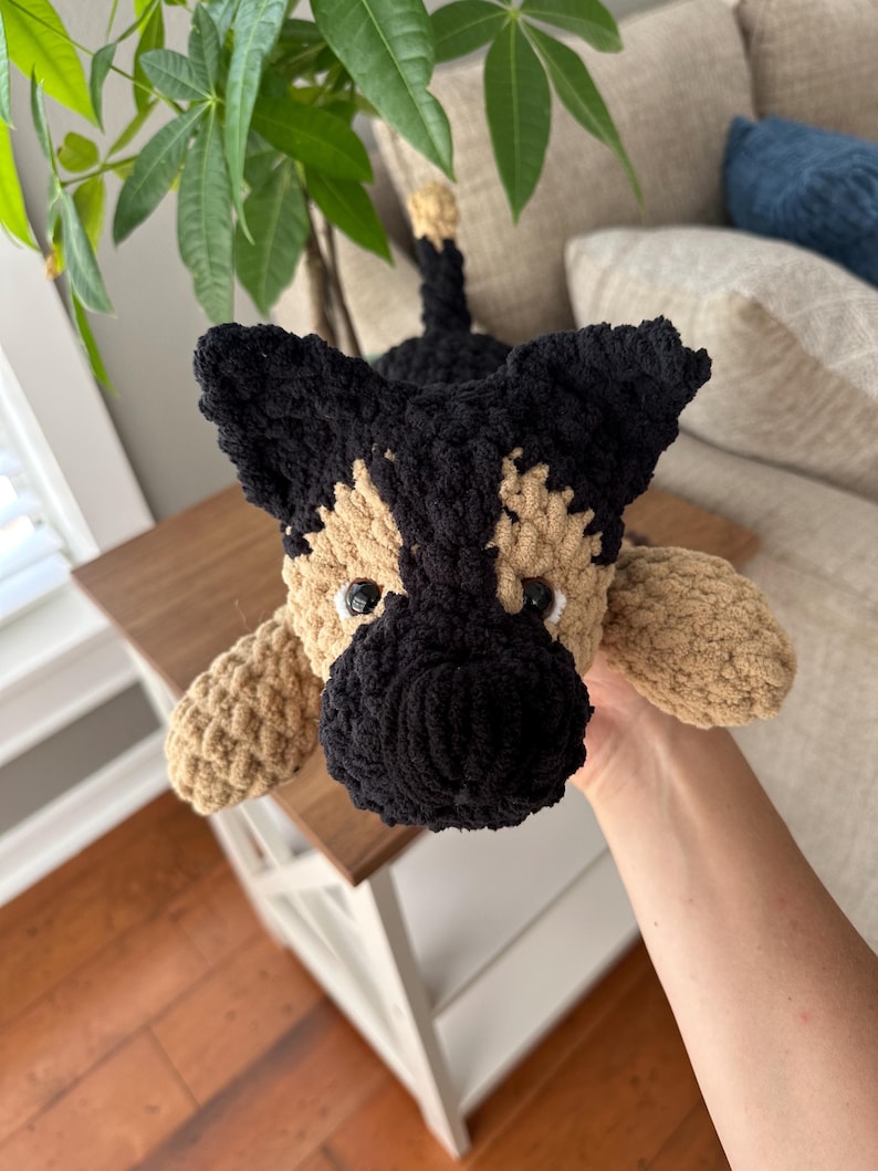 May include: A handmade crochet dog toy in the style of a German Shepherd. The dog has a black snout, ears, and back, with tan and cream-colored accents. The toy is lying down, with its paws extended.