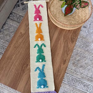 May include: A crocheted wall hanging with a fringe border features five colourful bunny silhouettes in pink, orange, green, and blue. The bunnies are arranged vertically in a row.