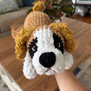 May include: Handmade crochet dog toy in the likeness of a Saint Bernard. The dog has a white face with black accents around the eyes and nose, and tan ears. The toy is made of soft, plush yarn.