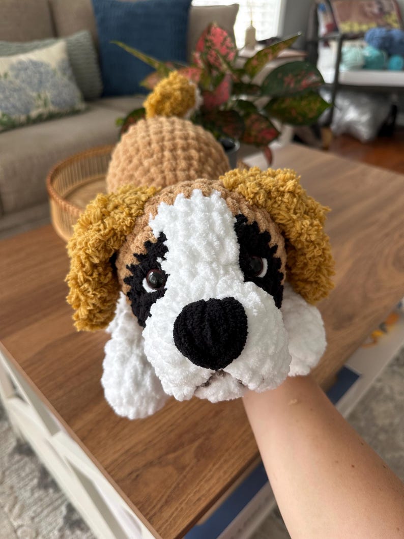 May include: Handmade crochet dog toy in the likeness of a Saint Bernard. The dog has a white face with black accents around the eyes and nose, and tan ears. The toy is made of soft, plush yarn.