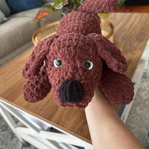 May include: A handmade, crocheted dog plushie in a rich, reddish-brown color. The dog has floppy ears, a black nose, and green eyes. The plush toy is soft and cuddly, perfect for children or as a decorative item.