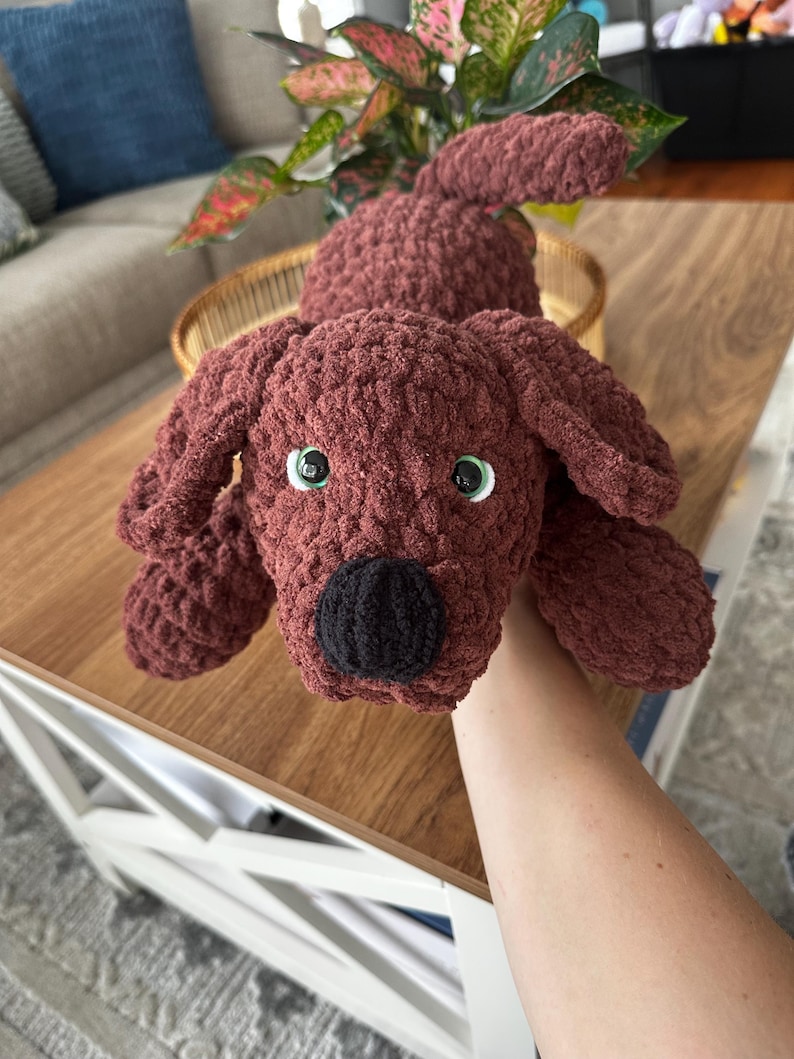 May include: A handmade, crocheted dog plushie in a rich, reddish-brown color. The dog has floppy ears, a black nose, and green eyes. The plush toy is soft and cuddly, perfect for children or as a decorative item.