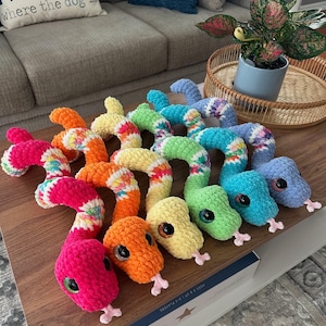 May include: A selection of brightly coloured, crocheted snake toys in shades of pink, orange, yellow, green, and blue. Each snake has large, googly eyes and a small pink tongue. The toys are displayed on a wooden table.