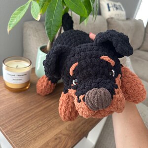 May include: A handmade crochet dog toy in black and tan. The plush toy has a detailed face with small eyes and a brown nose. The dog is lying down on a wooden surface.