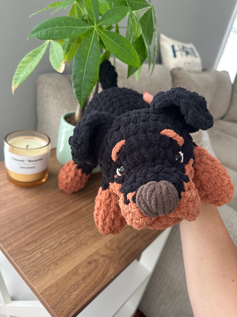 May include: A handmade crochet dog toy in black and tan. The plush toy has a detailed face with small eyes and a brown nose. The dog is lying down on a wooden surface.