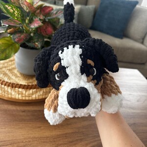 May include: A handmade crochet Bernese Mountain Dog plush toy. The dog has a black and white head, black ears, and brown and white paws. The plush is lying on a wooden table.
