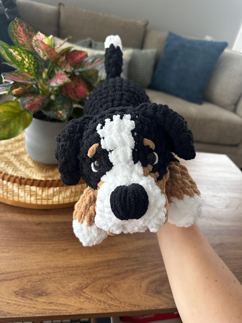 May include: A handmade crochet Bernese Mountain Dog plush toy. The dog has a black and white head, black ears, and brown and white paws. The plush is lying on a wooden table.