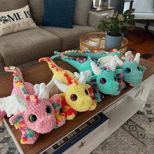 May include: A collection of five colorful, crocheted dragon plush toys. Each dragon features a unique color scheme, including pink, yellow, and teal, with rainbow accents and large, expressive eyes. They are arranged on a wooden coffee table.