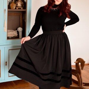 May include: A black long-sleeved turtleneck sweater paired with a high-waisted black midi skirt featuring two horizontal velvet bands near the hem. The skirt has a gathered waistline and a full silhouette, complemented by black lace-up boots.