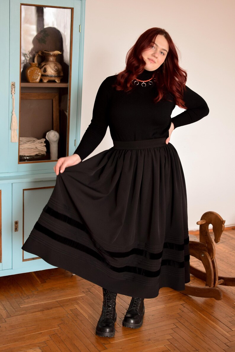 May include: A black long-sleeved turtleneck sweater paired with a high-waisted black midi skirt featuring two horizontal velvet bands near the hem. The skirt has a gathered waistline and a full silhouette, complemented by black lace-up boots.