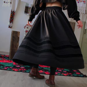 May include: A black skirt with three horizontal velvet bands, paired with a matching cropped top. The skirt is full and flowing. Handmade folk maxi skirt inspired by traditional European clothing, including Hungarian, Ukrainian, Romanian, Slovak, and Eastern European folk skirts. A versatile piece for boho maxi skirt looks, folk festival outfits, heritage-inspired fashion, slow fashion wardrobes, and handmade ethnic clothing.