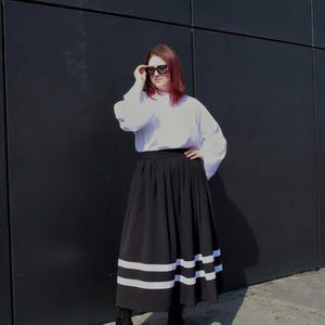 May include: A long black skirt featuring two horizontal white stripes near the hem, paired with a white long-sleeved top and black boots. The skirt has a gathered waistline and a full silhouette, while the top has loose sleeves and a high neckline.