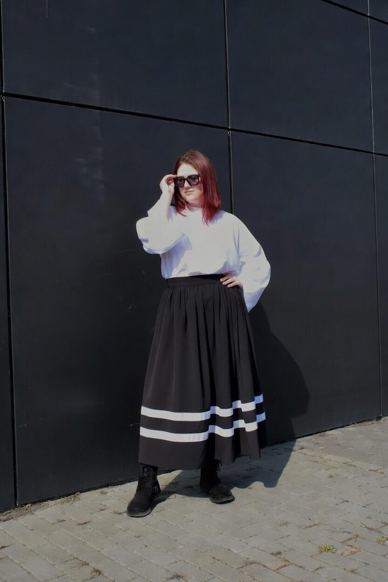May include: A long black skirt featuring two horizontal white stripes near the hem, paired with a white long-sleeved top and black boots. The skirt has a gathered waistline and a full silhouette, while the top has loose sleeves and a high neckline.