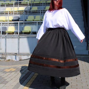 May include: A long black skirt featuring horizontal pleats and two wide brown satin ribbon bands near the hem, paired with a loose-fitting white long-sleeved top. The skirt has a full, flared silhouette and a high waistband, worn with dark footwear.