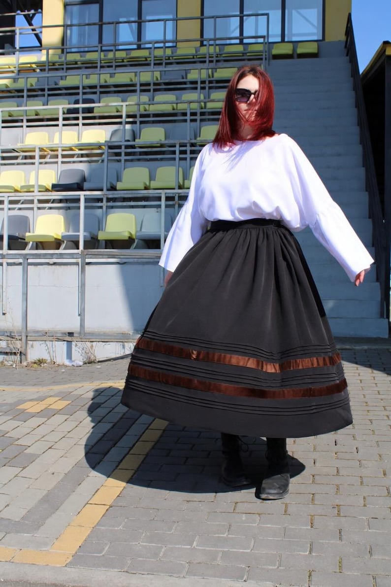May include: A long black skirt featuring horizontal pleats and two wide brown satin ribbon bands near the hem, paired with a loose-fitting white long-sleeved top. The skirt has a full, flared silhouette and a high waistband, worn with dark footwear.
