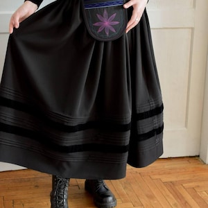 May include: A black midi skirt featuring a pleated waist, two horizontal velvet bands near the hem, and a detachable pouch at the front. The pouch is decorated with a purple and pink embroidered flower motif and blue velvet trim accents.